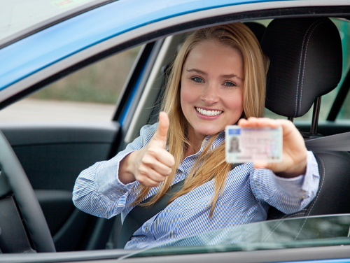 Teen driver in the driver seat of the car holds drivers license in one hand and gives thumbs up with the other hand